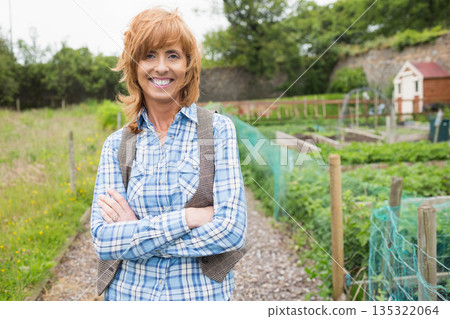 Mature woman standing on gravel path by raised beds with netting wearing plaid shirt, tweed vest 135322064