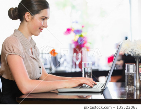 Female in twenties typing on silver laptop at cafe, water glass, white flower vase, copy space 135322065