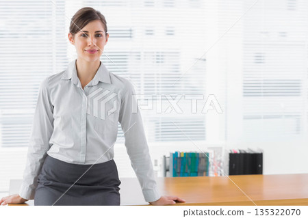 Professional woman leaning on desk facing camera in office with blinds and bookshelf, copy space 135322070