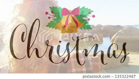Standing woman in red-black coat holding smartphone on sandy beach, with Christmas bells graphic Standing woman in red-black coat holding smartphone on sandy beach, with Christmas bells graphic 135322487