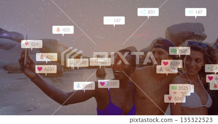 Smiling four friends posing for group selfie on sandy beach at sunset with smartphone notifications 135322523