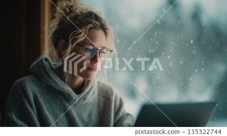 Focused woman working on laptop by rainy window creating cozy home office atmosphere 135322744