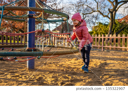 Little girl climbing rotating rope swing playground equipment in city park during spring. Outdoor childhood play, physical activity and everyday life in urban public space Little girl climbing rotating rope swing playground equipment in city park during spring. Outdoor childhood play, physical activity and everyday life in urban public space 135322907