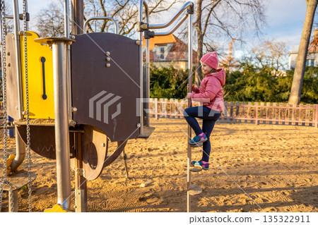 Little girl climbing playground structures in city park during spring. Childhood outdoor activity, physical play and everyday leisure moment in real life urban environment Little girl climbing playground structures in city park during spring. Childhood outdoor activity, physical play and everyday leisure moment in real life urban environment 135322911