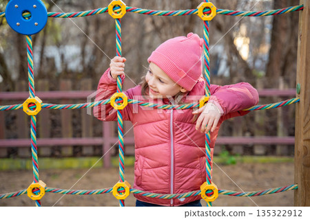 Little girl climbing playground structures in city park during spring. Childhood outdoor activity, physical play and everyday leisure moment in real life urban environment Little girl climbing playground structures in city park during spring. Childhood outdoor activity, physical play and everyday leisure moment in real life urban environment 135322912