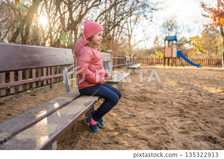 Little girl sitting on bench at playground in city park during spring. Outdoor childhood rest, calm moment and everyday life in urban public space Little girl sitting on bench at playground in city park during spring. Outdoor childhood rest, calm moment and everyday life in urban public space 135322913