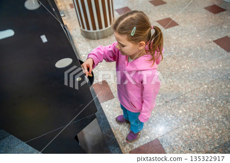Little girl collecting coins from money exchange machine in shopping mall kids area. Preparation for play, childhood independence moment and family leisure routine in real life commercial environment 135322917