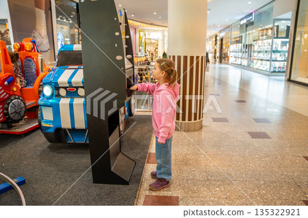 Little girl collecting coins from money exchange machine in shopping mall kids area. Preparation for play, childhood independence moment and family leisure routine in real life commercial environment 135322921