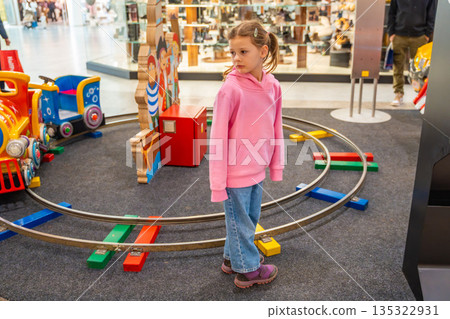 Little girl riding children train attraction in shopping mall kids area. Childhood entertainment activity, playful movement and family leisure time in real life commercial environment 135322931
