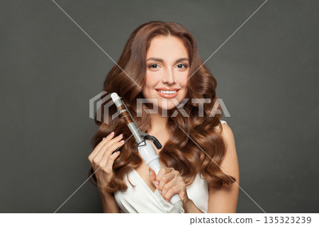 Glamorous woman with long wavy hairstyle using hair curling tongs iron, indoor portrait.  135323239