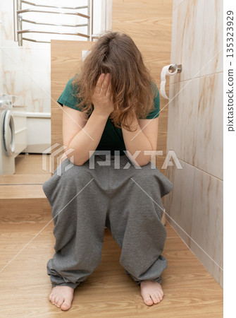 Woman sitting on toilet covering her face with hands, experiencing stomach discomfort and constipation in a bathroom. Authentic scene from life, personal space, daily routine, realistic life. Woman sitting on toilet covering her face with hands, experiencing stomach discomfort and constipation in a bathroom. Authentic scene from life, personal space, daily routine, realistic life. 135323929