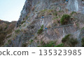 A low-angle detailed view of a steep rocky cliff face reinforced with a heavy-duty steel wire mesh netting designed to prevent rockfalls and landslides. 135323985