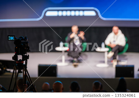 Panel discussion on stage with two speakers and camera recording audience at event conference Panel discussion on stage with two speakers and camera recording audience at event conference 135324061