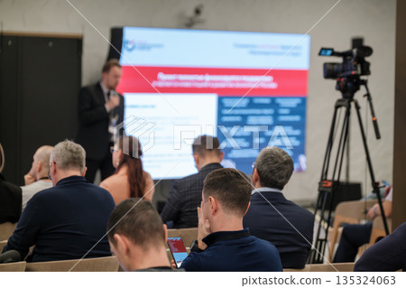 Business conference presentation with audience listening to a speaker on stage during a live event Business conference presentation with audience listening to a speaker on stage during a live event 135324063
