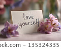 Macro shot of handwritten love yourself on card, soft-focus flowers in background, gentle pastel background, natural soft light 135324335