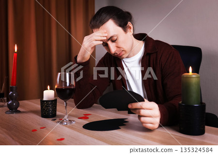 Sad man holding a broken black heart at a table with wine and candles. Upset lonely guy suffering from breakup or divorce. Anti-Valentine's Day concept. High quality photo 135324584