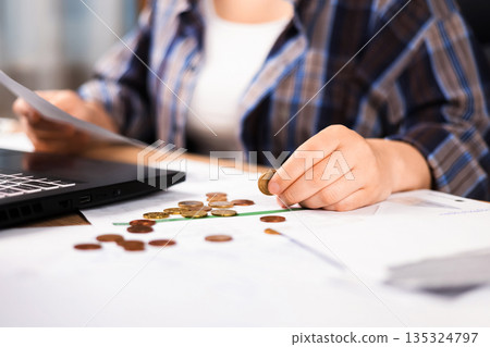 A woman holds a coin and checks accounts next to a laptop. Concept of family budget planning: money and documents are scattered across a wooden table. High quality photo A woman holds a coin and checks accounts next to a laptop. Concept of family budget planning: money and documents are scattered across a wooden table. High quality photo 135324797