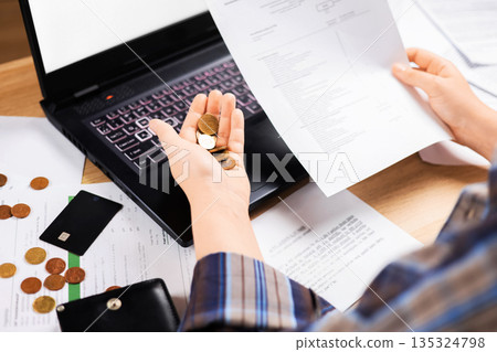 A woman holds euro coins and checks a banknote next to a laptop. Family budget concept: money, a credit card, and documents on a wooden table. High quality photo A woman holds euro coins and checks a banknote next to a laptop. Family budget concept: money, a credit card, and documents on a wooden table. High quality photo 135324798