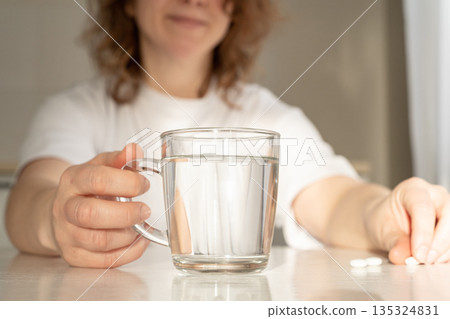 A woman holds a glass of water, preparing to take white pills lying on a table. Focus on health and well-being. Daily medication and healthcare concept 135324831