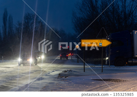Night winter road Magdeburg heavy snowfall cars truck driving icy junction illuminated headlights streetlight. Direction sign low visibility snow covered asphalt traffic danger transport challenge 135325985