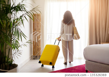 Rear view of adult woman pulling yellow suitcase toward bright hotel window Rear view of adult woman pulling yellow suitcase toward bright hotel window 135326369