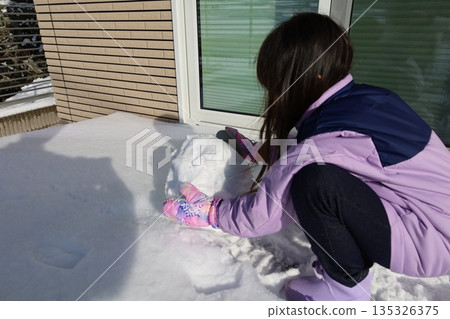 Girl making a snowman, enjoying winter moments in front of her house 135326375