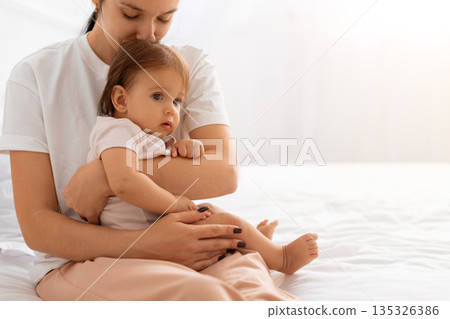 Caucasian mother holding baby close while sitting on bed, showing emotional connection 135326386