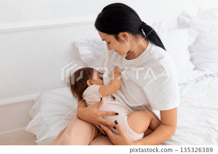 Young caucasian mother gently breastfeeding her infant on white bed at home 135326389