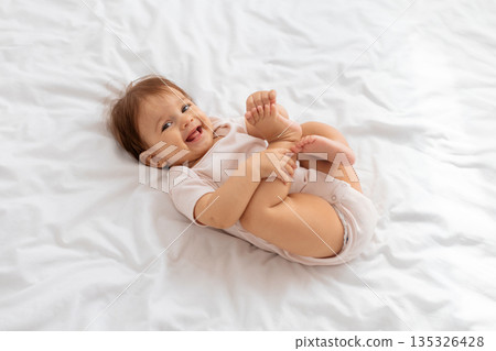 Cheerful infant baby relaxing on white bed in bright room Cheerful infant baby relaxing on white bed in bright room 135326428
