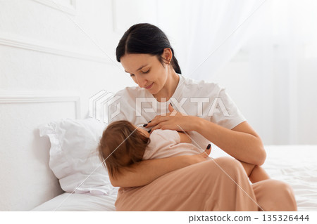 Mother breastfeeding her baby while sitting on bed in home interior 135326444