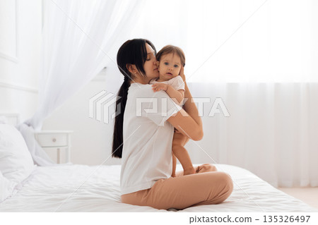 Young mother gently holding her baby while sitting on white bed 135326497