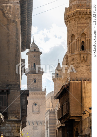 Historic Qalawun Complex in Cairo, featuring intricate architecture, tall minarets, and domes under a blue sky with clouds. A significant cultural landmark on Al-Muizz Street. Historic Qalawun Complex in Cairo, featuring intricate architecture, tall minarets, and domes under a blue sky with clouds. A significant cultural landmark on Al-Muizz Street. 135327116