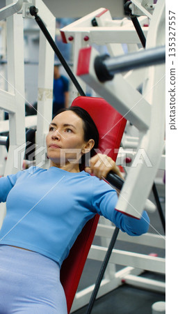 Gym workout. Bench press in hammer machine. Caucasian woman trains her chest muscles with focused bench press reps on the hammer machine during an intense gym workout. Gym workout. Bench press in hammer machine. Caucasian woman trains her chest muscles with focused bench press reps on the hammer machine during an intense gym workout. 135327557