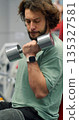 Gym workout. Close-up vertical photo of a fit bearded man training biceps with dumbbells during an intense gym workout. Seated biceps with dumbbells workout. 135327581