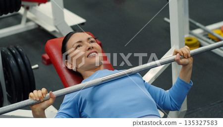 Gym workout. Bench press. Caucasian woman trains her pectoral muscles in the gym. Caucasian woman trains chest muscles using bench press during gym workout. Close-up photo. 135327583