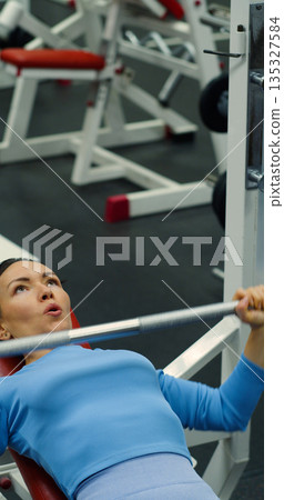 Gym workout. Bench press. Caucasian woman trains her pectoral muscles in the gym. Caucasian woman trains chest muscles using bench press during gym workout. Gym workout. Bench press. Caucasian woman trains her pectoral muscles in the gym. Caucasian woman trains chest muscles using bench press during gym workout. 135327584