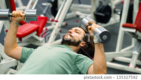 A bearded Caucasian man performs dumbbell bench presses, focusing on chest strength in detailed close-up photo. Intense workout targeting chest muscles by a focused gym-goer. 135327592