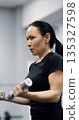 Close-up vertical photo of a fit middle aged woman training biceps with dumbbells during an intense gym workout. Biceps with dumbbells workout. Cropped footage of a working out woman. 135327598