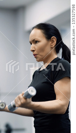 Close-up vertical photo of a fit middle aged woman training biceps with dumbbells during an intense gym workout. Biceps with dumbbells workout. Cropped footage of a working out woman. Close-up vertical photo of a fit middle aged woman training biceps with dumbbells during an intense gym workout. Biceps with dumbbells workout. Cropped footage of a working out woman. 135327598