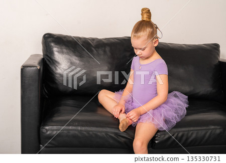 Young ballerina with a cochlear implant sits on a black couch during a quiet rehearsal break. Inclusive ballet, hearing technology, and focused childhood through rest and reflection. Young ballerina with a cochlear implant sits on a black couch during a quiet rehearsal break. Inclusive ballet, hearing technology, and focused childhood through rest and reflection. 135330731