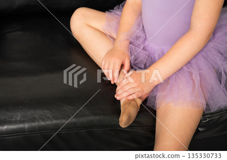A close view of a young dancer adjusting her ballet shoe on a sofa. Inclusive training, hearing support, and careful preparation through dance and discipline. 135330733