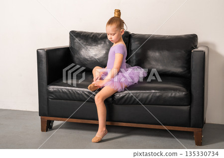 A young ballerina with a cochlear implant sits quietly on a black couch after rehearsal. Inclusive ballet, hearing technology, and focused childhood through rest and recovery. A young ballerina with a cochlear implant sits quietly on a black couch after rehearsal. Inclusive ballet, hearing technology, and focused childhood through rest and recovery. 135330734