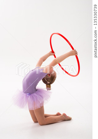 A young gymnast with a cochlear implant holds a red hoop in a graceful side pose. Inclusive gymnastics, hearing support, and physical coordination through rhythmic ballet training. A young gymnast with a cochlear implant holds a red hoop in a graceful side pose. Inclusive gymnastics, hearing support, and physical coordination through rhythmic ballet training. 135330739