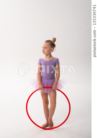 A smiling ballerina with a cochlear implant raises a red hoop in a ballet pose. Inclusive sport, hearing technology, and confident childhood expression through rhythmic dance and movement. A smiling ballerina with a cochlear implant raises a red hoop in a ballet pose. Inclusive sport, hearing technology, and confident childhood expression through rhythmic dance and movement. 135330741