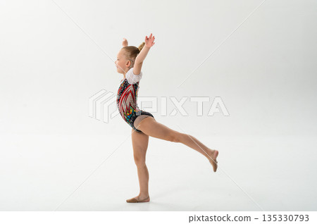 Young dancer with a cochlear implant performs a graceful arabesque in a white studio. Inclusive sport, hearing technology, and childhood confidence through rhythmic gymnastics and ballroom movement. 135330793