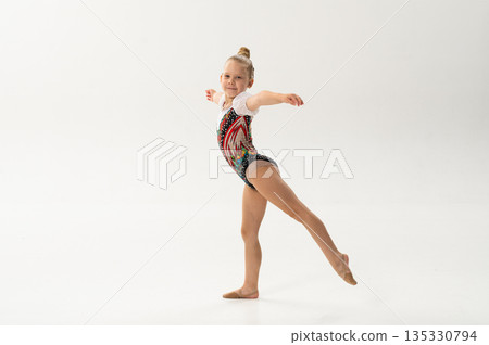Young dancer with a cochlear implant performs a graceful arabesque in a white studio. Inclusive sport, hearing technology, and childhood confidence through rhythmic gymnastics and ballroom movement. 135330794