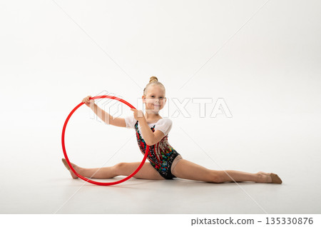 Young rhythmic gymnast with cochlear implant holding red hoop in studio. Hearing rehabilitation, physical flexibility and inclusive childhood sports through artistic training 135330876