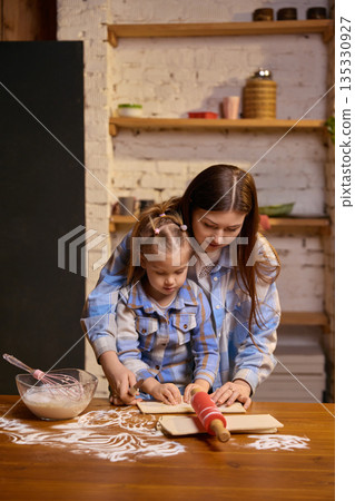 Mother guiding daughter rolling dough at wooden kitchen table. Mother guiding daughter rolling dough at wooden kitchen table. 135330927