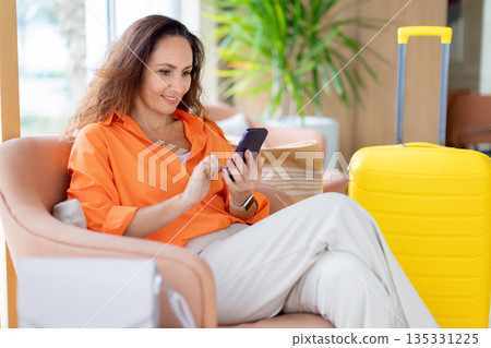 Adult caucasian woman sitting in hotel lounge chair using smartphone near suitcase Adult caucasian woman sitting in hotel lounge chair using smartphone near suitcase 135331225