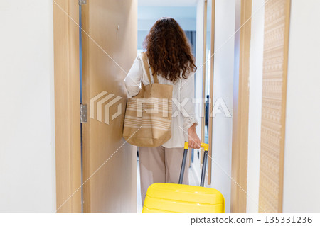 Rear view of adult caucasian woman entering hotel room pulling suitcase 135331236